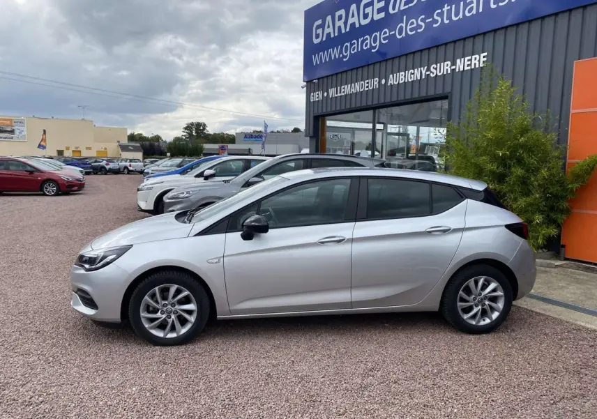 Profil côté gauche d'une Opel Astra 2021 Sovereign Silver avec jantes alliage 16 pouces, stationnée devant un garage.