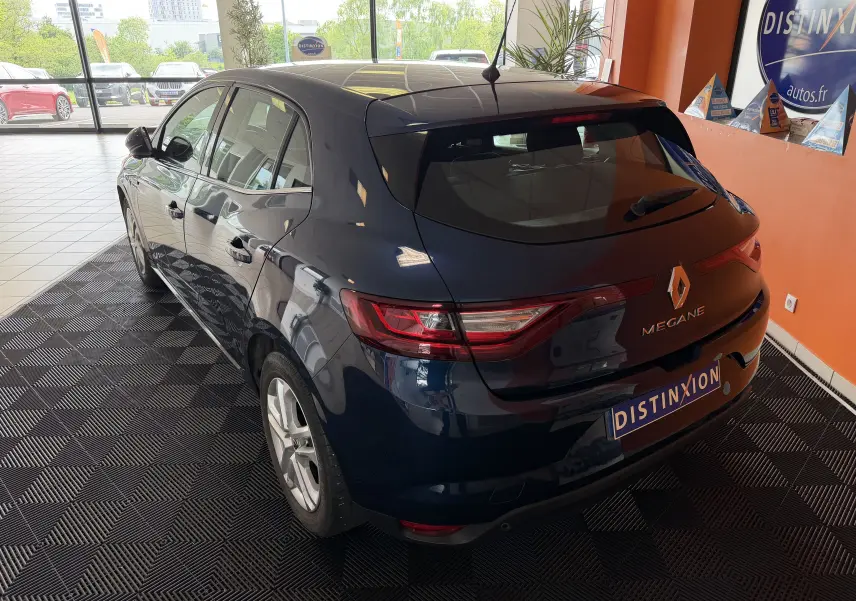 Vue 3/4 arrière droite d'une Renault Mégane bleu foncé 5 portes, en intérieur sur sol noir texturé, avec logo et feux arrière visibles.