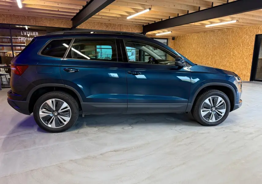 Vue de profil côté gauche d'un Skoda Karoq bleu 2023 avec jantes alu 17'' dans un garage en bois.