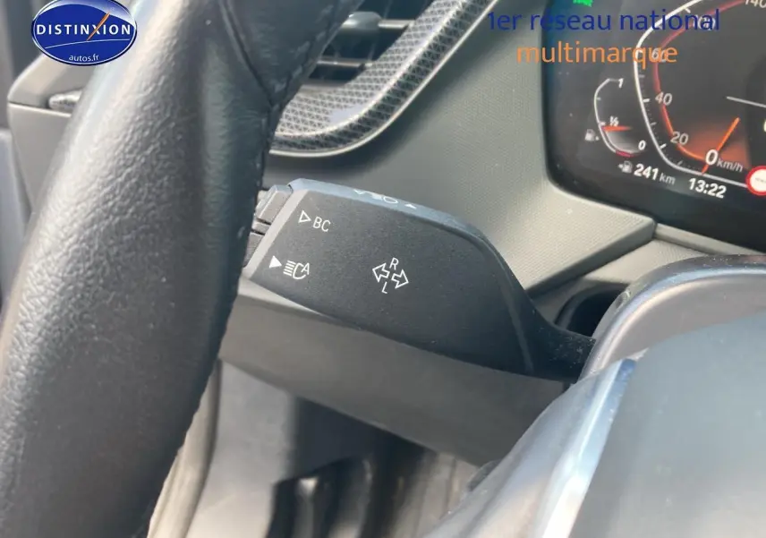 Gros plan sur le commodo gauche du volant d'une BMW Série 1 gris foncé, tableau de bord et compteur visibles en arrière-plan.