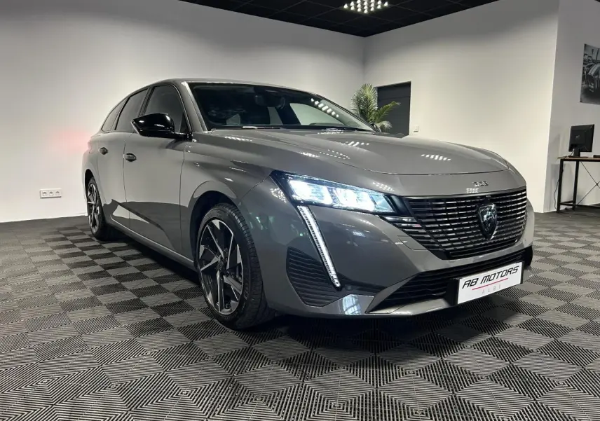 Peugeot 308 SW gris Sélénium vue 3/4 avant droit avec calandre noire et feux LED allumés en intérieur showroom.