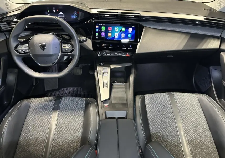 Vue intérieure avant de la Peugeot 308 SW 2025, tableau de bord noir avec écran tactile et sièges bi-matière gris et noir.