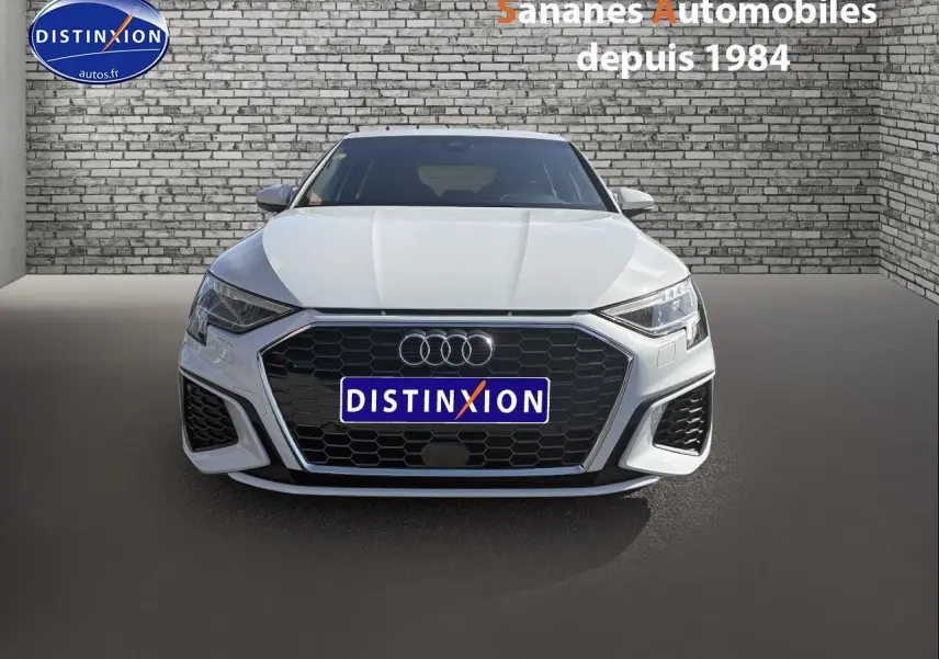 Vue frontale d'une Audi A3 Sportback blanche 2023 avec calandre noire et projecteurs LED, sur fond mural en briques.