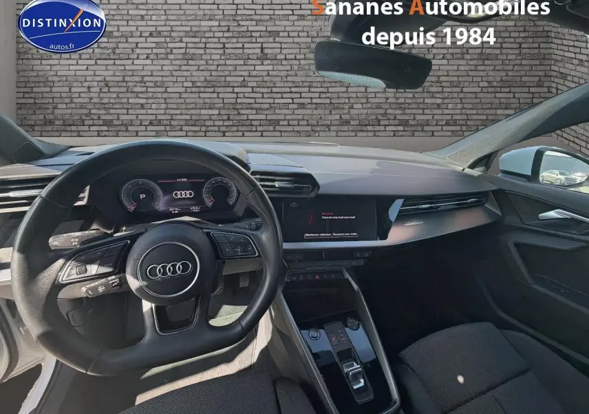 Vue intérieure avant de l'Audi A3 Sportback blanche 2023, mettant en valeur le volant cuir multifonctions et le tableau de bord digital.
