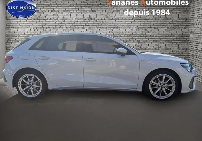 Profil droit d'une Audi A3 Sportback blanche 2023 avec jantes alliage et détails S line visibles sur fond de mur en briques.