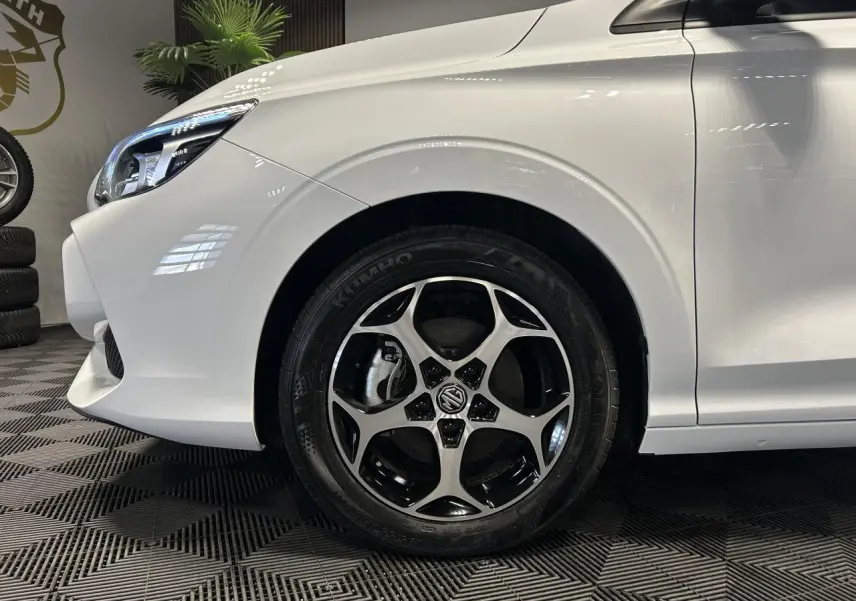 Gros plan sur la roue avant gauche et le phare d’un MG3 blanc 2025 dans un intérieur showroom.