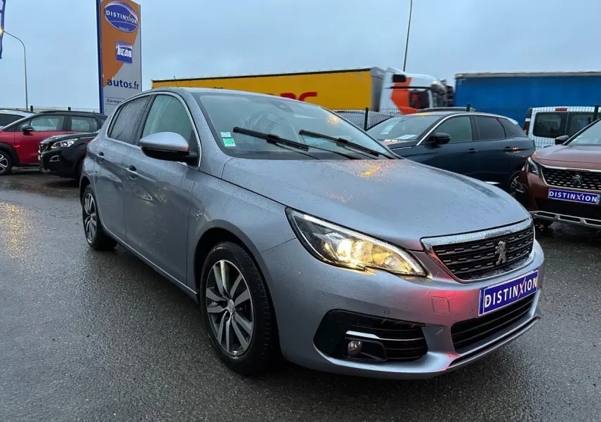 Peugeot 308 gris Artense vue 3/4 avant droit sous la pluie avec phares allumés sur parking extérieur.
