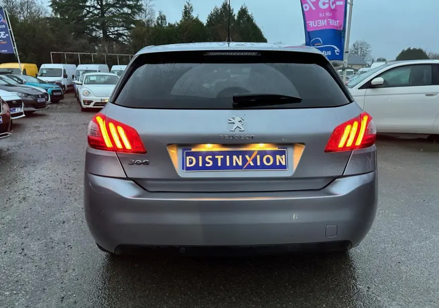 Vue arrière d'une Peugeot 308 gris Artense avec feux allumés sur un parking extérieur.