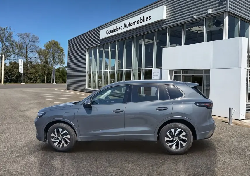 Vue latérale droite d'un Volkswagen Tiguan 1.5 eHybrid gris, avec jantes alliage Venezia 17 pouces, devant un bâtiment commercial.