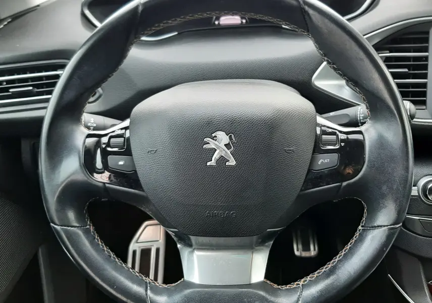 Vue rapprochée du volant en cuir noir du Peugeot 308 SW 2021, avec commandes intégrées et tableau de bord en arrière-plan.