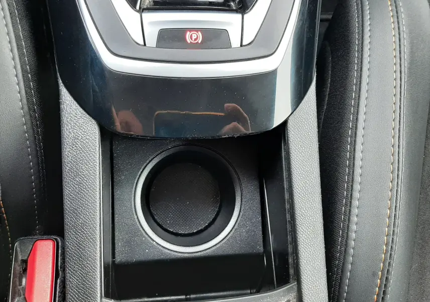 Vue rapprochée de la console centrale noire avec bouton de frein à main électrique et porte-gobelet, sièges tissu gris avec surpiqûres.