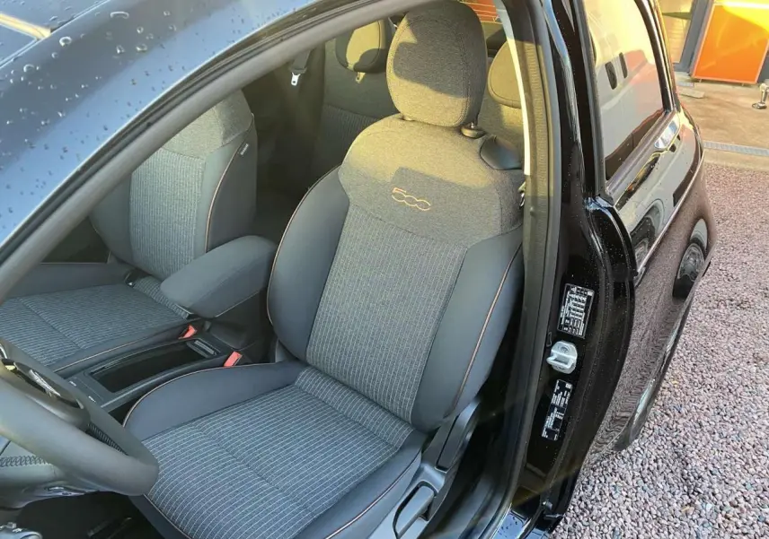 Vue intérieure côté conducteur d'une FIAT 500 électrique noire, siège avant gris avec logo 500 brodé et porte ouverte.