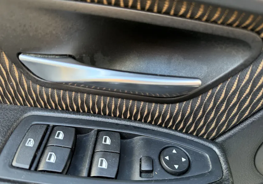 Gros plan sur la poignée intérieure et les commandes de vitres électriques côté conducteur d'une BMW Série 3 320D gris avec tissu rayé.