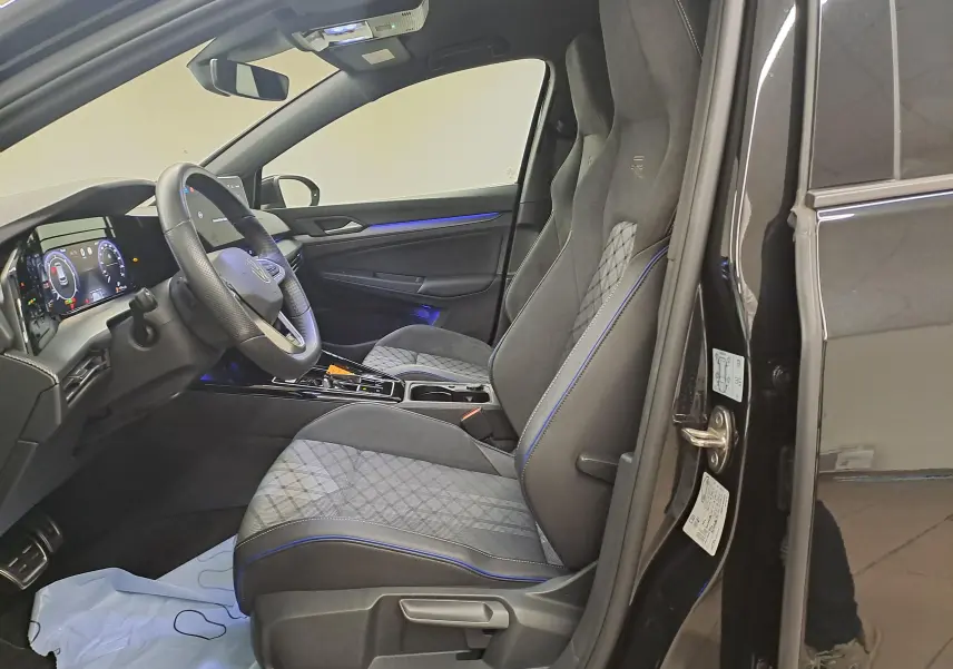 Vue intérieure côté conducteur de la Volkswagen Golf 2.0 TDI R Line 2024 noir grenadille avec sièges sport et éclairage d’ambiance bleu.