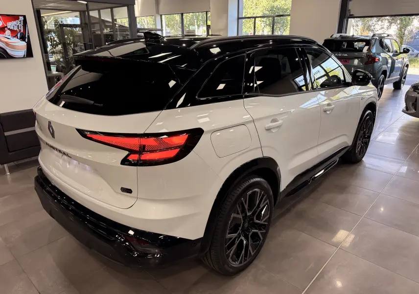 Vue 3/4 arrière droite du Renault Austral blanc nacré avec toit noir, jantes noires et feux arrière LED allumés en showroom.