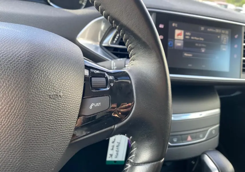 Gros plan sur le volant cuir noir de la Peugeot 308 gris aluminium, avec commandes intégrées et écran tactile en arrière-plan.