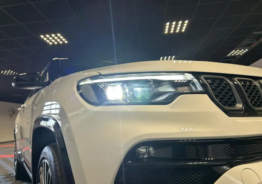 Vue 3/4 avant gauche d’un Jeep Compass blanc avec toit noir et phares LED allumés en intérieur showroom.