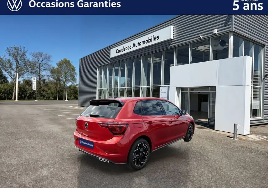 Vue 3/4 arrière droite d'une Volkswagen Polo rouge métallisée garée devant un bâtiment moderne de concession.