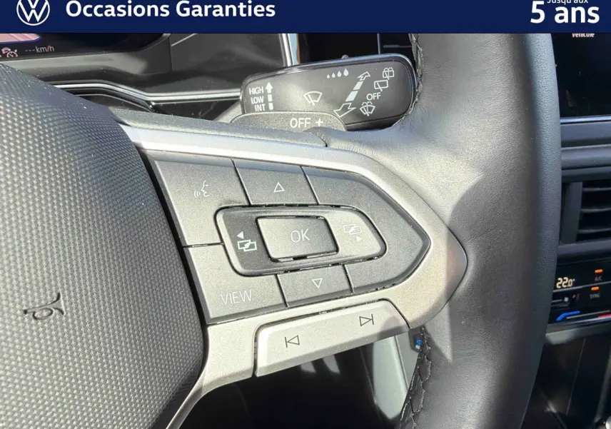 Gros plan sur les commandes multifonctions gauche du volant cuir du Volkswagen Taigo gris, avec tableau de bord et climatisation tactile en arrière-plan.