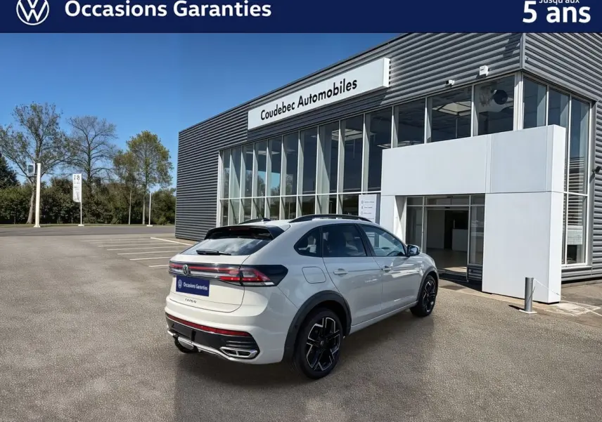 Vue 3/4 arrière droite d'un Volkswagen Taigo gris avec attelage amovible, devant un bâtiment de concession.