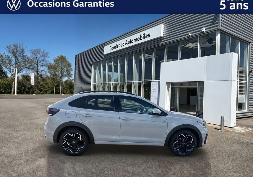 Vue latérale droite d'un Volkswagen Taigo gris devant un bâtiment moderne avec jantes alliage noires et détails noirs contrastants.