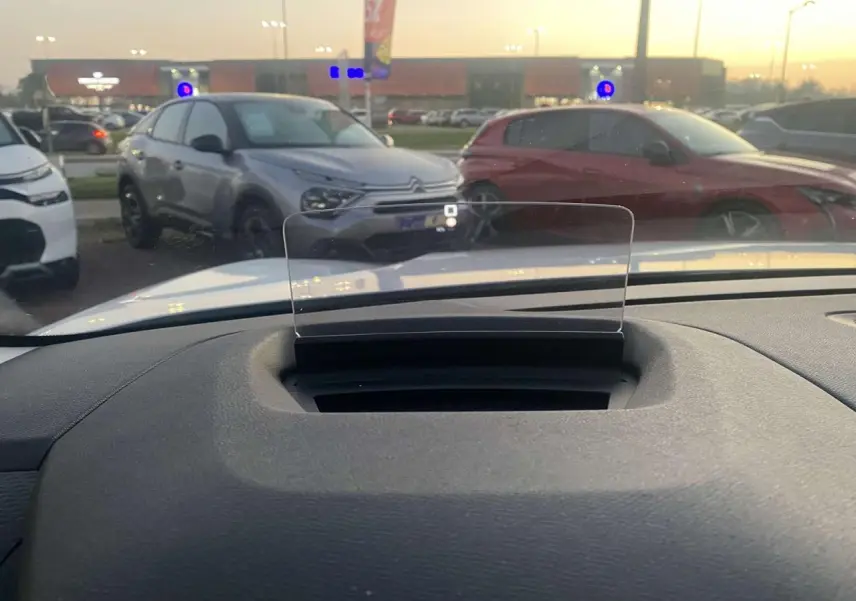 Vue depuis l'habitacle montrant le pare-brise et l'affichage tête haute d'une Citroën C4 blanche stationnée en extérieur au crépuscule.