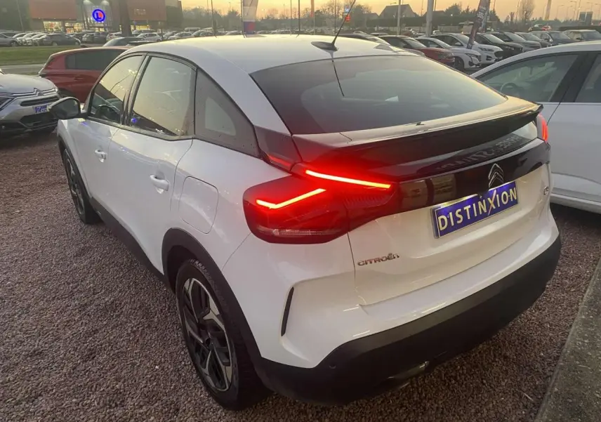 Vue 3/4 arrière droite d'une Citroën C4 blanche avec feux arrière allumés sur parking en soirée.