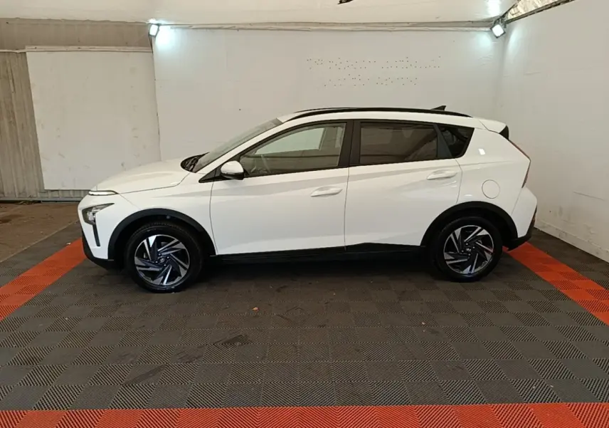 Vue de profil côté gauche d'un Hyundai Bayon blanc 2024 avec jantes noires et toit noir dans un garage.