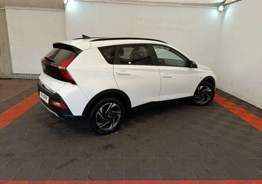 Vue 3/4 arrière droite d'une Hyundai Bayon blanche 2024 avec jantes noires et toit noir dans un garage.