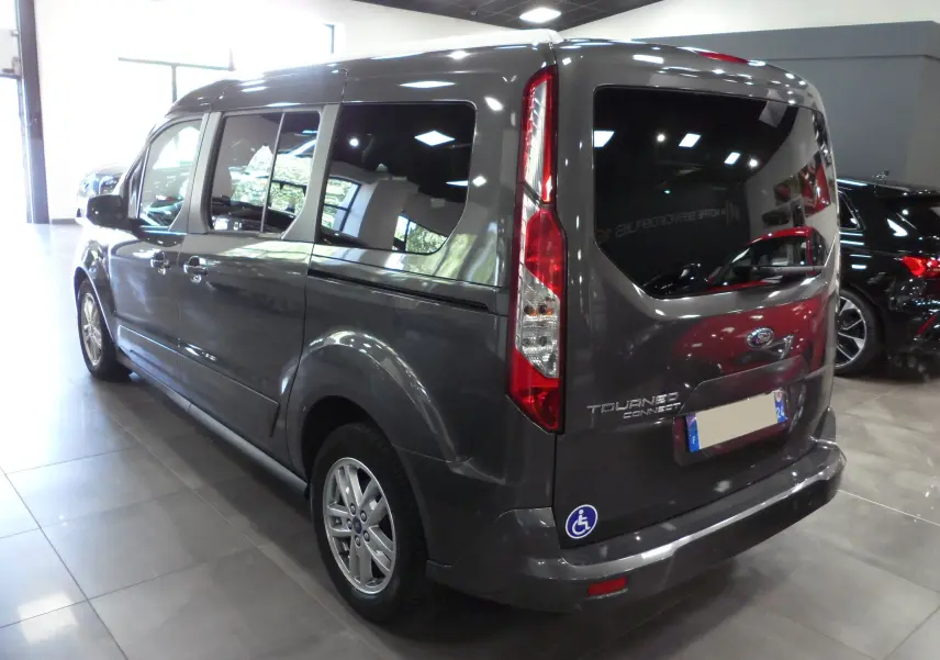 Vue 3/4 arrière droite du Ford Grand Tourneo Connect gris Magnetic avec autocollant handicapé sur le pare-chocs arrière.