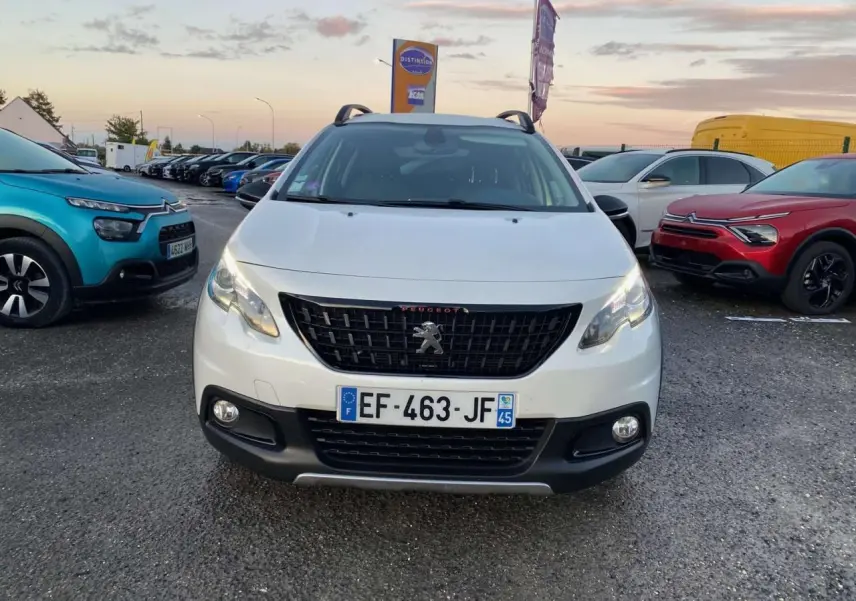 Peugeot 2008 blanc vue de face, avec calandre noire et phares allumés sur un parking extérieur.