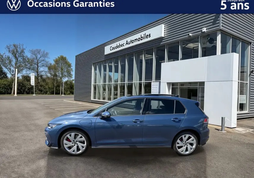 Vue de profil côté gauche d'une Volkswagen Golf 1.5 eHybrid 272 DSG6 bleu métallisé garée devant un bâtiment moderne.
