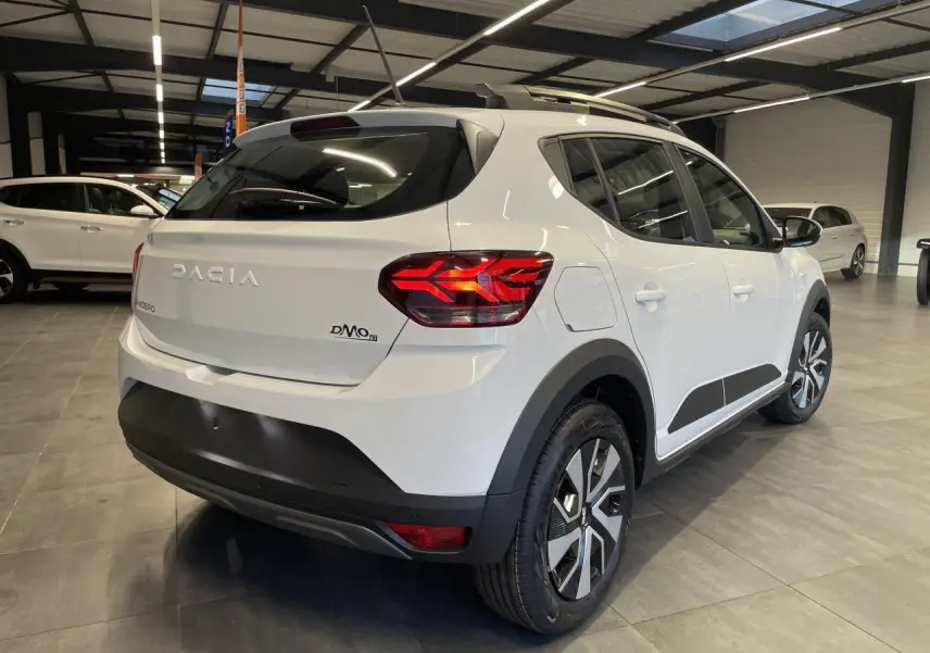 Vue 3/4 arrière droite d'une Dacia Sandero TCe 90 Stepway blanche avec barres de toit et jantes spécifiques en concession.