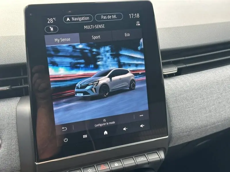 Écran tactile intérieur montrant une Renault Clio blanche en mouvement de profil droit sur fond urbain flou.