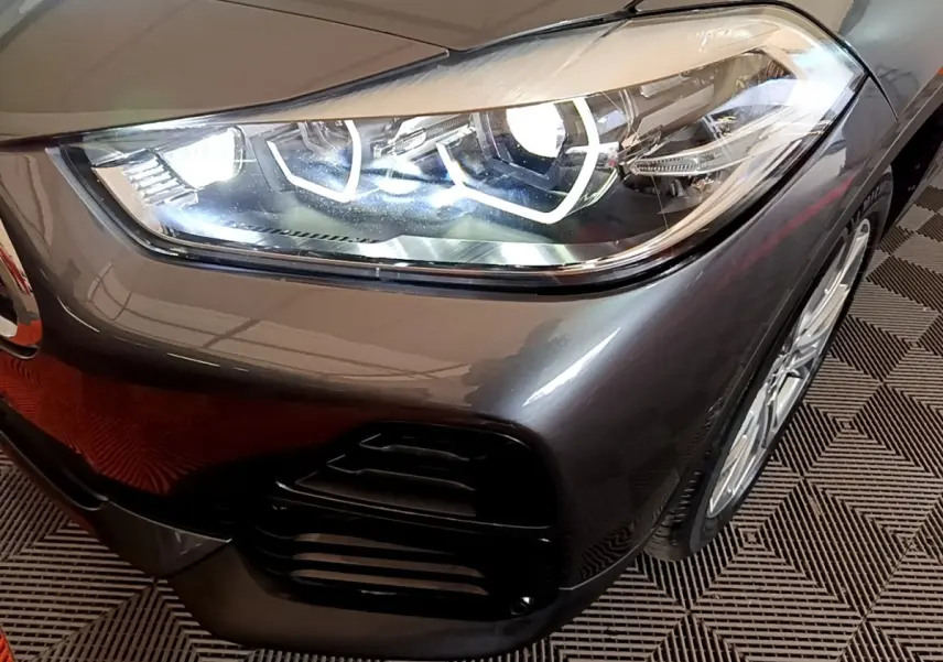 Gros plan sur le phare avant droit allumé du BMW X2 xDrive25e gris métallisé avec jante visible sur sol à motif.