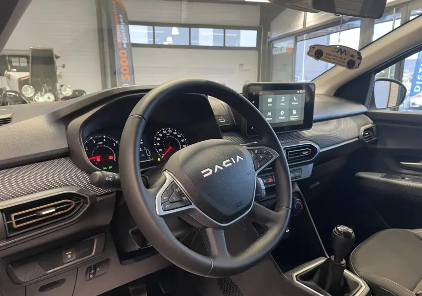 Intérieur avant droit d’une Dacia Sandero blanche 2025, volant avec logo, écran tactile et levier de vitesses manuel.