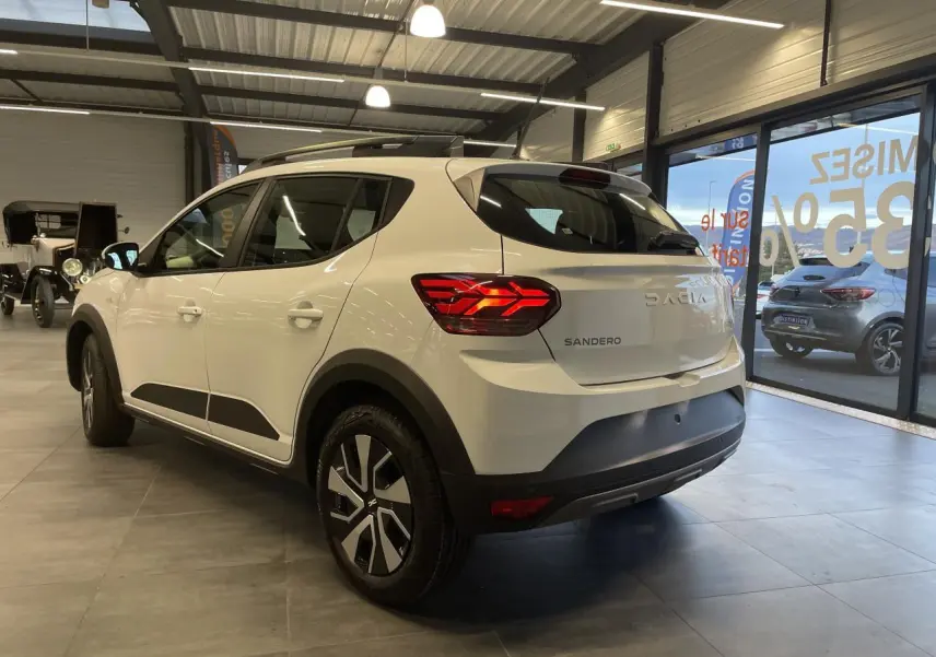 Vue 3/4 arrière droite d'une Dacia Sandero Stepway blanche glacier avec barres de toit et feux arrière LED en intérieur showroom.