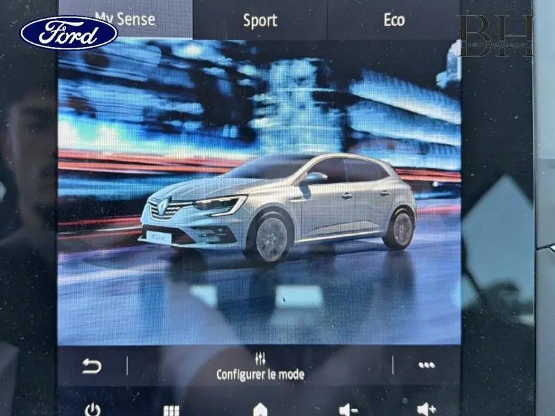 Vue 3/4 avant d'une Renault Mégane gris Highland affichée sur écran multimédia avec arrière-plan urbain flouté.