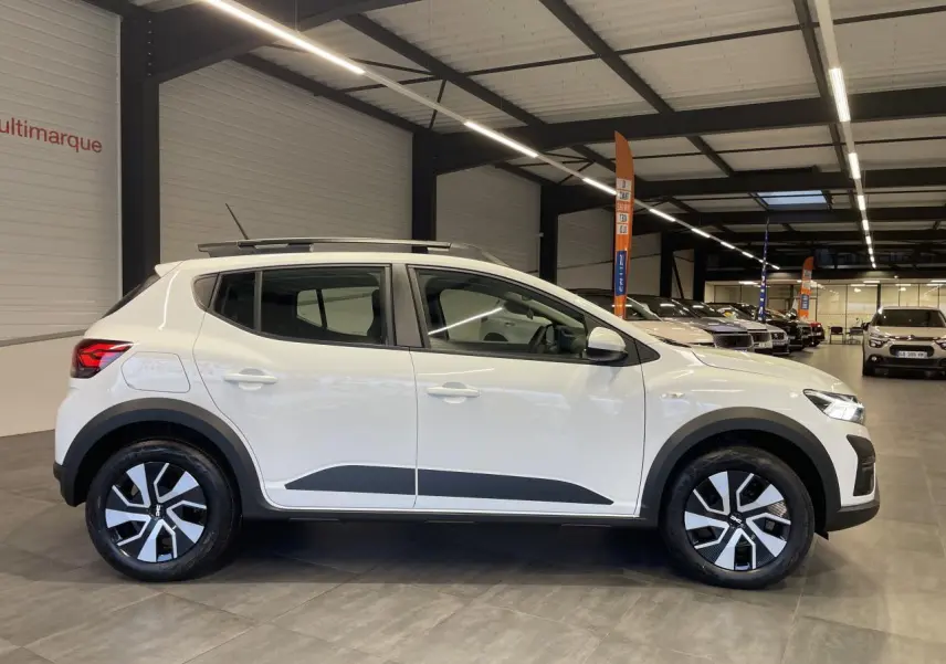 Vue de profil côté gauche d'une Dacia Sandero Stepway 2025 blanc glacier avec roues spécifiques et roue de secours visible en intérieur showroom.