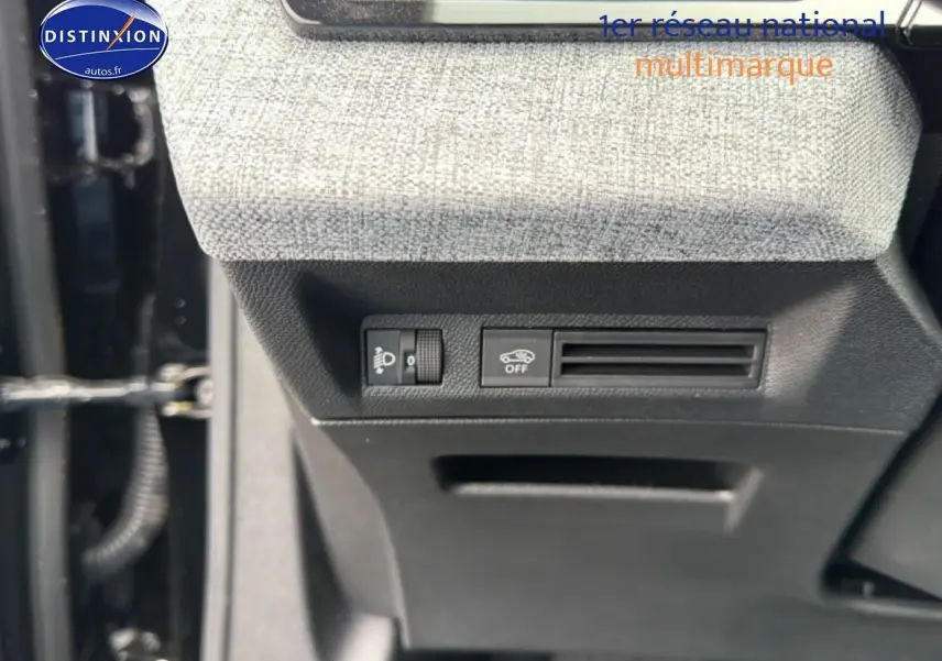 Détail intérieur montrant les commandes d'éclairage et de désactivation de l'assistance au freinage sur tissu gris et plastique noir.