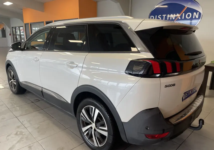 Vue 3/4 arrière droit d'un Peugeot 5008 blanc nacré avec feux arrière à griffes et attelage visible en intérieur showroom.