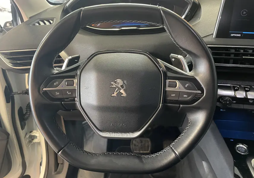 Vue rapprochée du volant cuir noir avec logo Peugeot d'un 5008 blanc nacré, tableau de bord et commandes visibles.