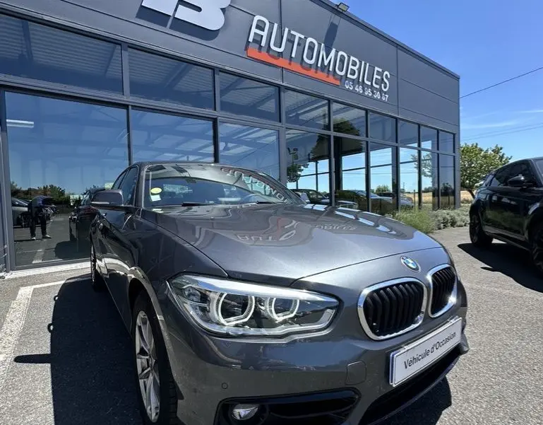 BMW Série 1 gris foncé vue en 3/4 avant droit devant un concessionnaire FB Automobiles par temps ensoleillé.