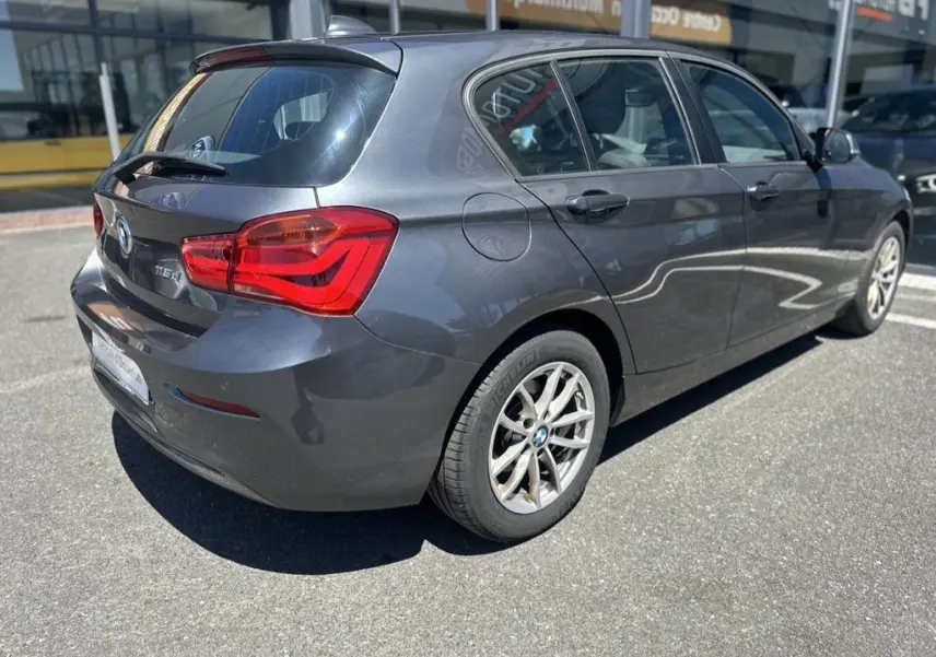 BMW Série 1 F20 gris foncé vue 3/4 arrière droit, mettant en valeur les feux arrière et les jantes alliage.