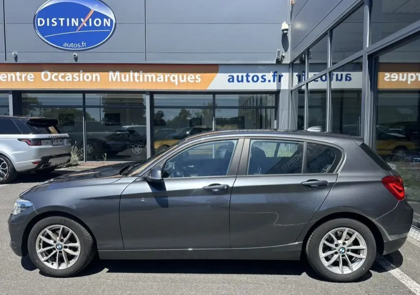 Profil côté gauche d'une BMW Série 1 F20 gris foncé, modèle 116D Business Design 2019, garée devant un showroom.