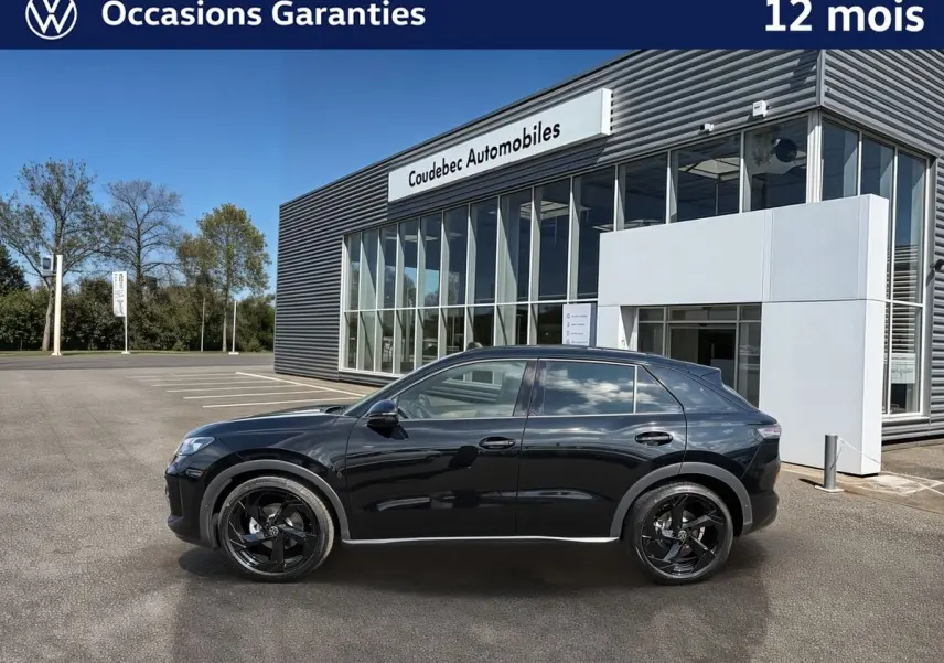 Profil droit d'un Volkswagen T-Roc noir métallisé avec jantes noires, garé devant un bâtiment moderne de concession.