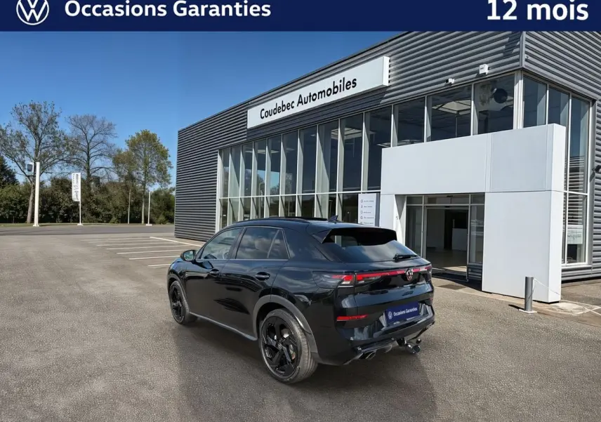 Volkswagen T-Roc noir en 3/4 arrière, avec jantes noires et crochet d'attelage, devant un bâtiment moderne.