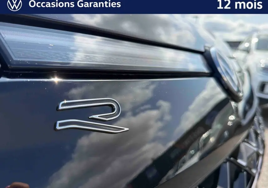 Gros plan sur l'avant noir brillant d'un Volkswagen T-Roc avec logo "R" et calandre détaillée en reflet.