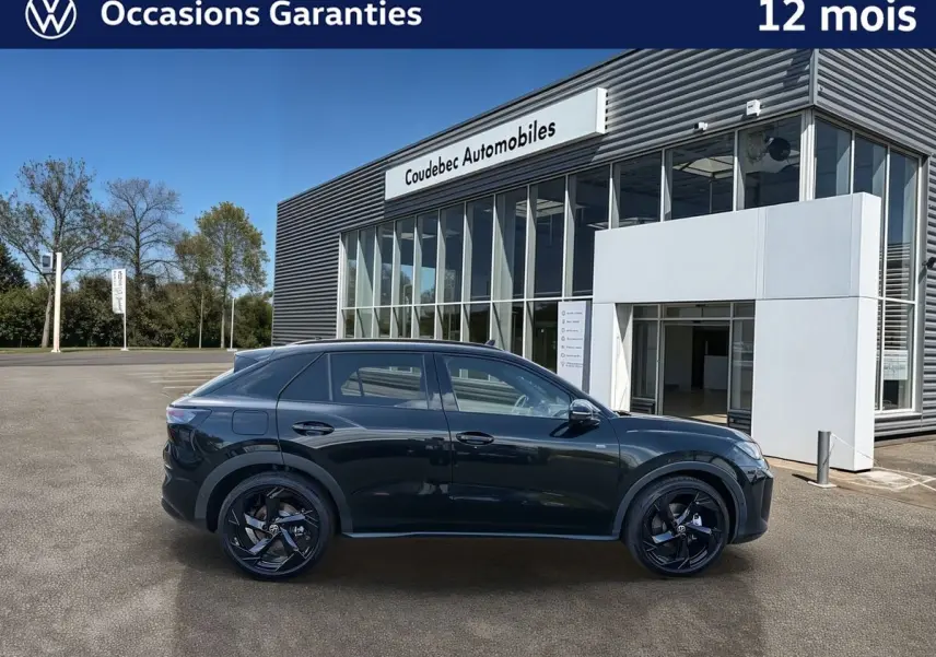 Vue de profil côté gauche d’un Volkswagen T-Roc noir métallisé avec jantes noires devant un bâtiment de concession.
