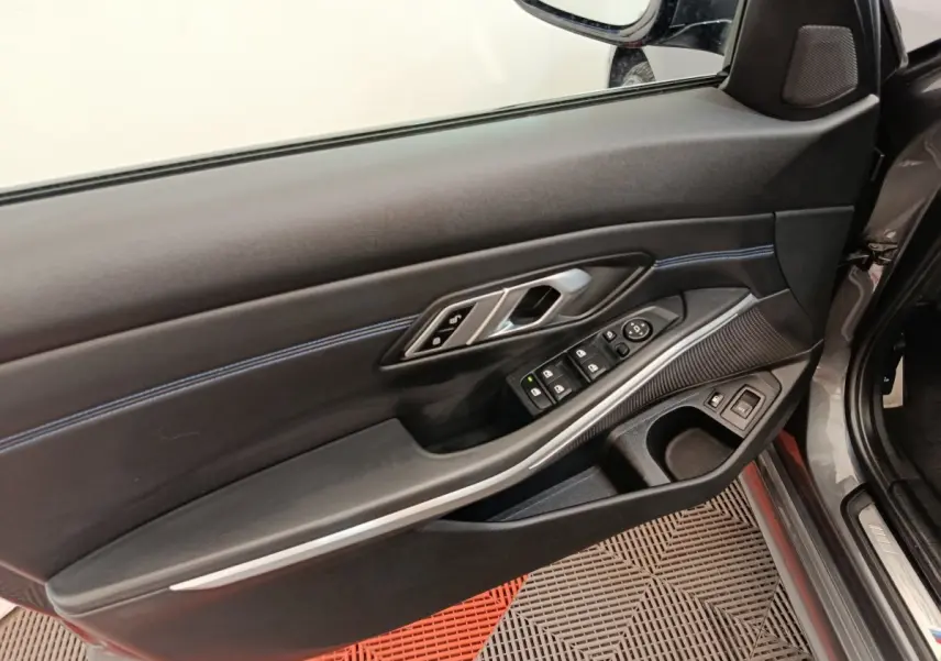 Vue intérieure côté conducteur de la porte d’une BMW Série 3 320e gris foncé avec commandes électriques et surpiqûres bleues.